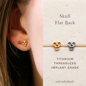 May include: A gold-colored skull-shaped stud earring and a small gold ball stud earring. The text reads: Skull Flat Back, TITANIUM THREADLESS IMPLANT GRADE. A gold and a silver-colored skull stud are shown separately on a textured surface.