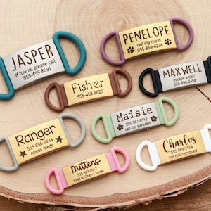 May include: A selection of personalised pet ID tags in colours such as teal, brown, pink, and green. Each tag displays a name, phone number, and extra text like "needs meds" or "call my mom". The tags are made of metal and silicone.