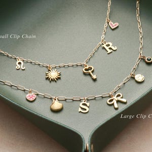 May include: Two gold-colored clip chain necklaces with various charms, including a sun, key, heart, letter, and bow. The necklaces are displayed on a green surface. The text "Small Clip Chain" and "Large Clip Chain" are visible.
