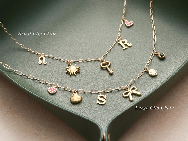 May include: Two gold-colored clip chain necklaces with various charms, including a sun, key, heart, letter, and bow. The necklaces are displayed on a green surface. The text "Small Clip Chain" and "Large Clip Chain" are visible.