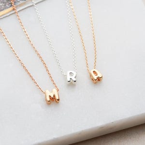 May include: Three delicate gold necklaces with initial charms. The charms are in the shape of the letters M, R, and A. The necklaces are on a white surface.