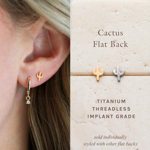 May include: Gold and silver cactus-shaped earrings. One earring is worn on the ear, and two are displayed on a beige surface. The text reads "Cactus Flat Back" and "TITANIUM THREADLESS IMPLANT GRADE."