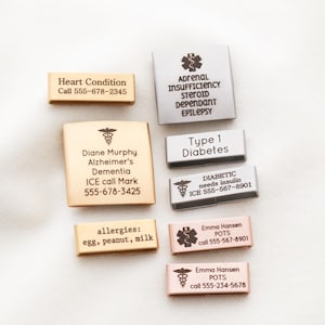 May include: A collection of rectangular medical alert watch band charms in gold, silver, and rose gold tones. Each charm features engraved medical information, including names, conditions like diabetes or epilepsy, and emergency contact phone numbers.