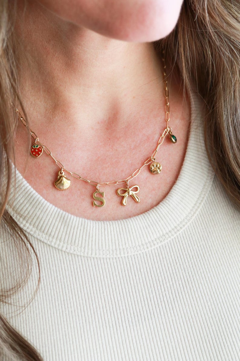 May include: Gold-toned charm necklace with a variety of pendants, including a strawberry, seashell, letter "S", bow, paw print, and a green gem. The necklace is worn on a person wearing a cream-colored shirt.