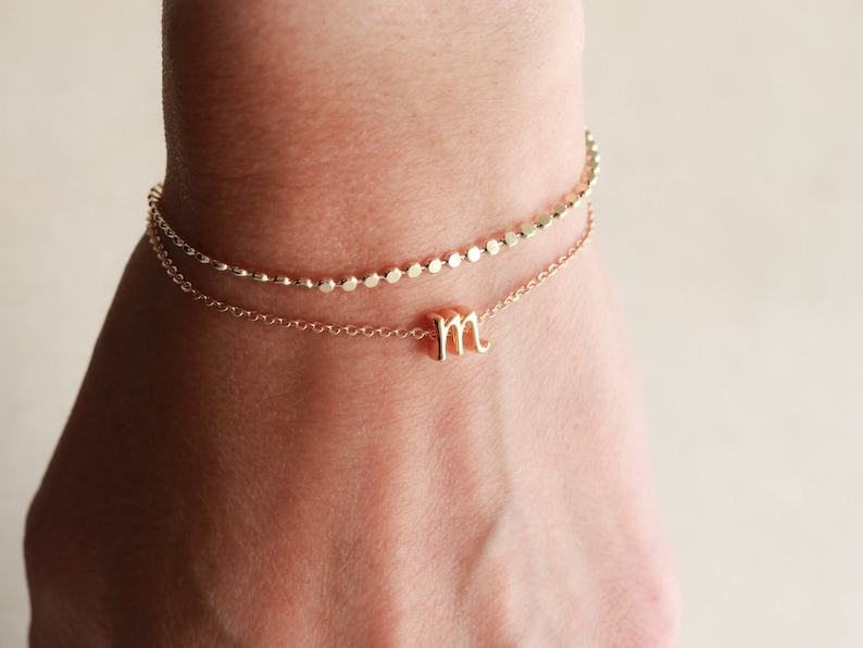 May include: A gold chain bracelet with a small letter "M" charm. The bracelet is layered with a second chain with small round beads.