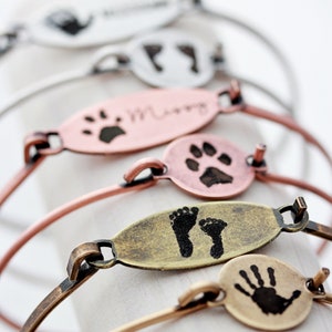May include: A collection of metal bangle bracelets featuring oval and circular charms. The charms are finished in silver, copper, and brass tones, each engraved with black ink impressions of handprints, footprints, or paw prints, with some including names.