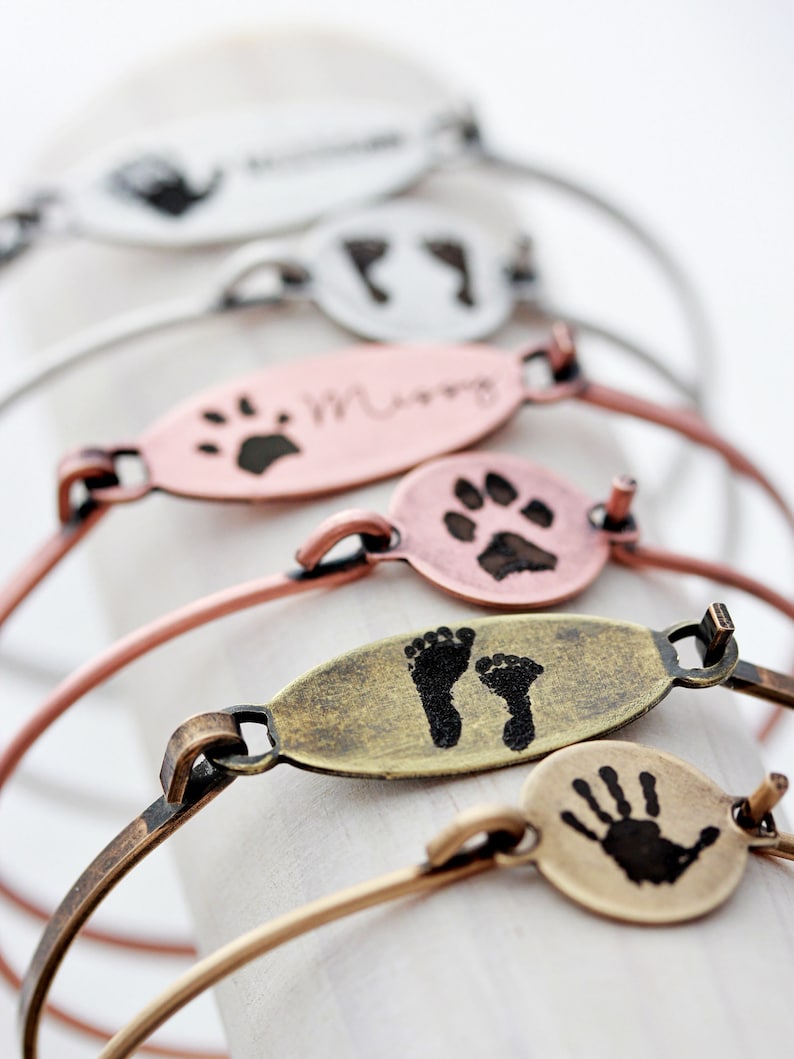 May include: A collection of metal bangle bracelets featuring oval and circular charms. The charms are finished in silver, copper, and brass tones, each engraved with black ink impressions of handprints, footprints, or paw prints, with some including names.