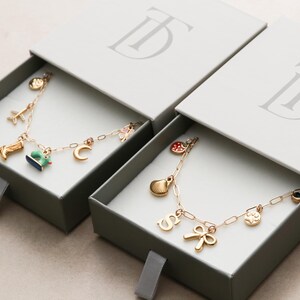 May include: Two gold charm bracelets with various charms, including a boot, airplane, and crescent moon, presented in gray boxes with the brand's logo. The bracelets are displayed in open boxes.