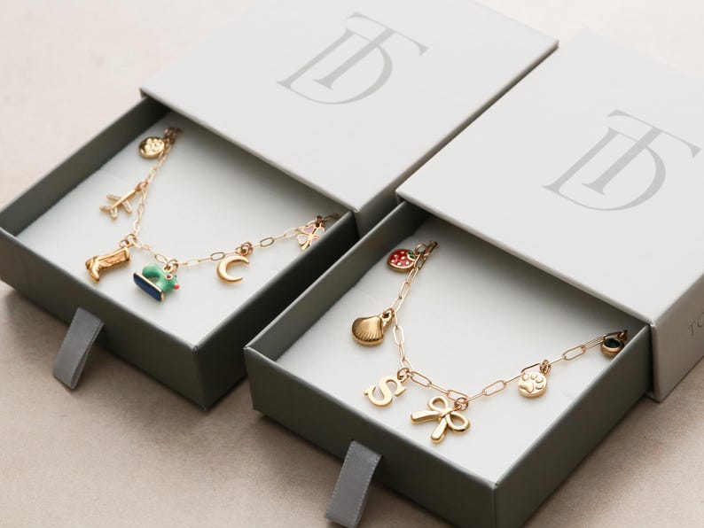 May include: Two gold charm bracelets with various charms, including a boot, airplane, and crescent moon, presented in gray boxes with the brand's logo. The bracelets are displayed in open boxes.