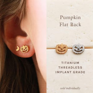 May include: A gold-toned pumpkin-shaped stud earring with a flat back, alongside a small crescent moon stud. The text reads Pumpkin Flat Back, Titanium Threadless Implant Grade. The design features a carved jack-o'-lantern face with cutouts.