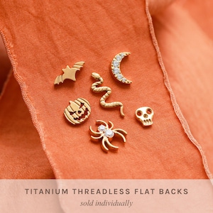 May include: A collection of gold-toned titanium threadless flat back earrings featuring various shapes including a bat, a crescent moon with clear stones, a snake, a jack-o'-lantern, a skull, and a spider with clear stones, labeled Titanium Threadless Flat Backs.