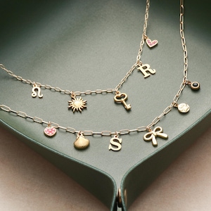May include: Two gold-colored necklaces with various charms, including a sun, key, bow, and letters. One necklace has a strawberry charm. The necklaces are displayed on a dark green tray.