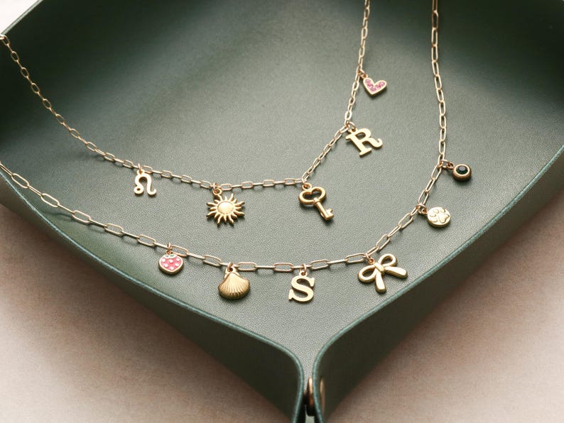 May include: Two gold-colored necklaces with various charms, including a sun, key, bow, and letters. One necklace has a strawberry charm. The necklaces are displayed on a dark green tray.
