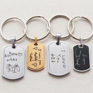 May include: Four metal keychains with rectangular tags featuring engraved handwriting and drawings. Two tags are silver-toned, one is gold-toned, and one is black with a silver border. Each tag is attached to a circular silver split ring.