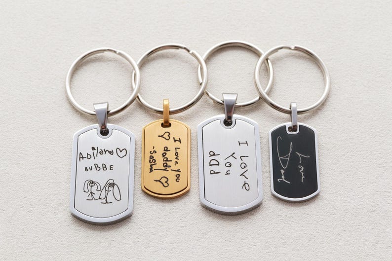May include: Four metal keychains with rectangular tags featuring engraved handwriting and drawings. Two tags are silver-toned, one is gold-toned, and one is black with a silver border. Each tag is attached to a circular silver split ring.