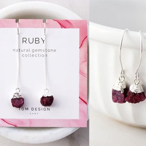 May include: Silver drop earrings featuring raw ruby gemstones. The earrings have a natural, organic look with a silver cap on each ruby. The card behind the earrings reads "RUBY natural gemstone collection" and "TOM DESIGN SHOP".