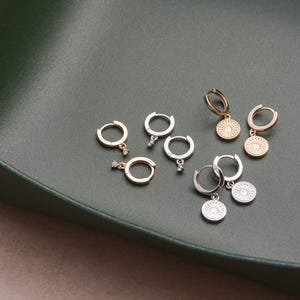 May include: A set of six gold and silver hoop earrings with a small dangling charm. Each charm is a circular design with a raised center and a small clear stone.