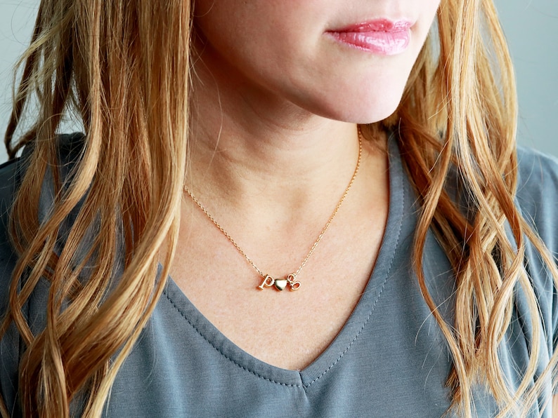 May include: A gold necklace with a heart charm and the letters "P" and "S" hanging from it. The necklace is worn by a person with long blonde hair.