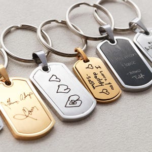May include: A collection of rectangular metal keychains with rounded corners, featuring custom laser-engraved handwriting and drawings. The keychains come in gold and silver tones, each attached to a circular metal split ring for keys.