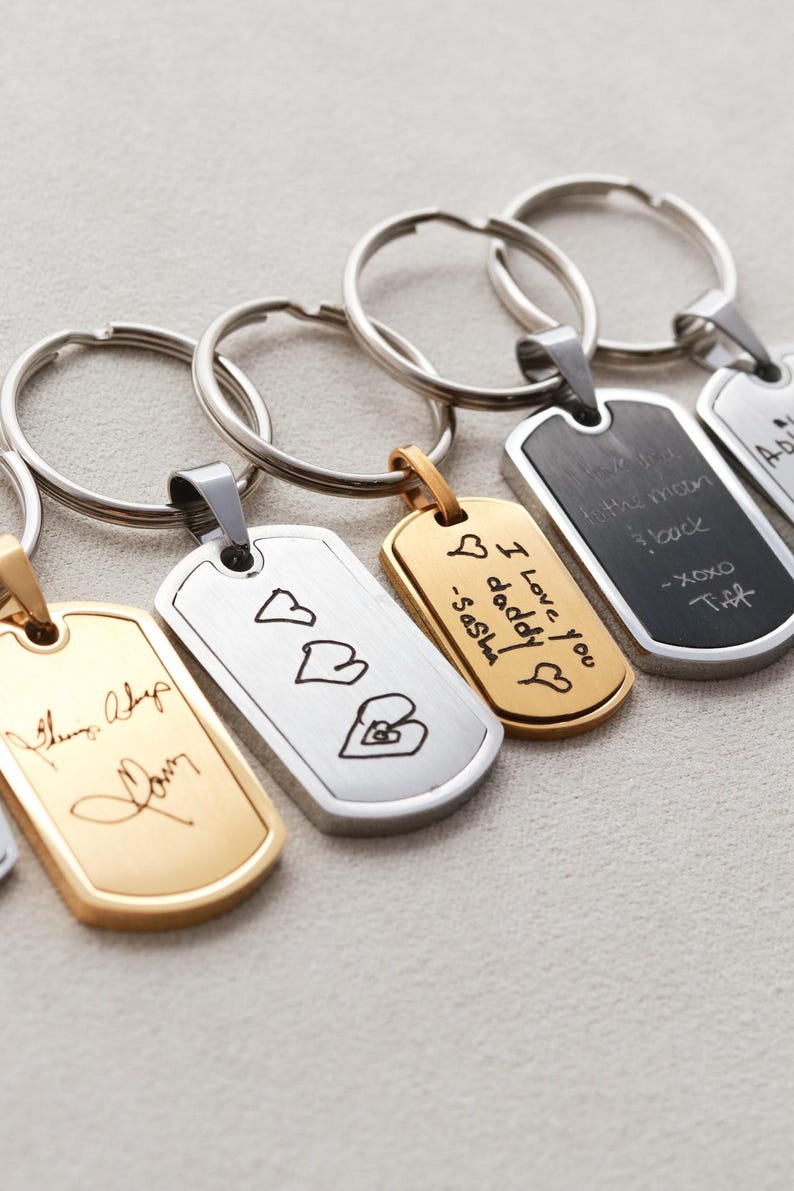 May include: A collection of rectangular metal keychains with rounded corners, featuring custom laser-engraved handwriting and drawings. The keychains come in gold and silver tones, each attached to a circular metal split ring for keys.