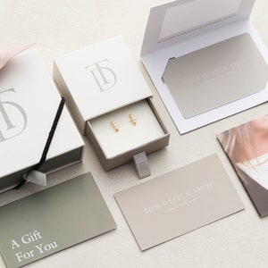 May include: A white gift box with a drawer containing a pair of gold earrings. The box has the initials "TD" embossed on the top. The gift box is surrounded by other gift packaging items, including a card that says "A Gift For You" and a card that says "TOM DESIGN SHOP".