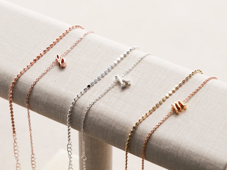 May include: Three delicate necklaces with initial charms. The necklaces are made of silver and rose gold chains with small round beads. The charms are in the shape of the letters 'E', 'K', and 'M'.