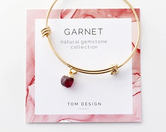May include: Gold-tone adjustable bracelet with a raw, dark red garnet gemstone pendant. The card behind the bracelet reads "GARNET natural gemstone collection" and "TOM DESIGN SHOP". The background has a pink watercolor design.