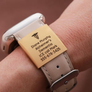 May include: A light beige watch band featuring a rectangular gold-toned metal medical alert tag engraved with a caduceus symbol and the text: Diane Murphy, Alzheimer's, Dementia, ICE call Mark, 555-678-3425. The band has a silver-toned buckle.