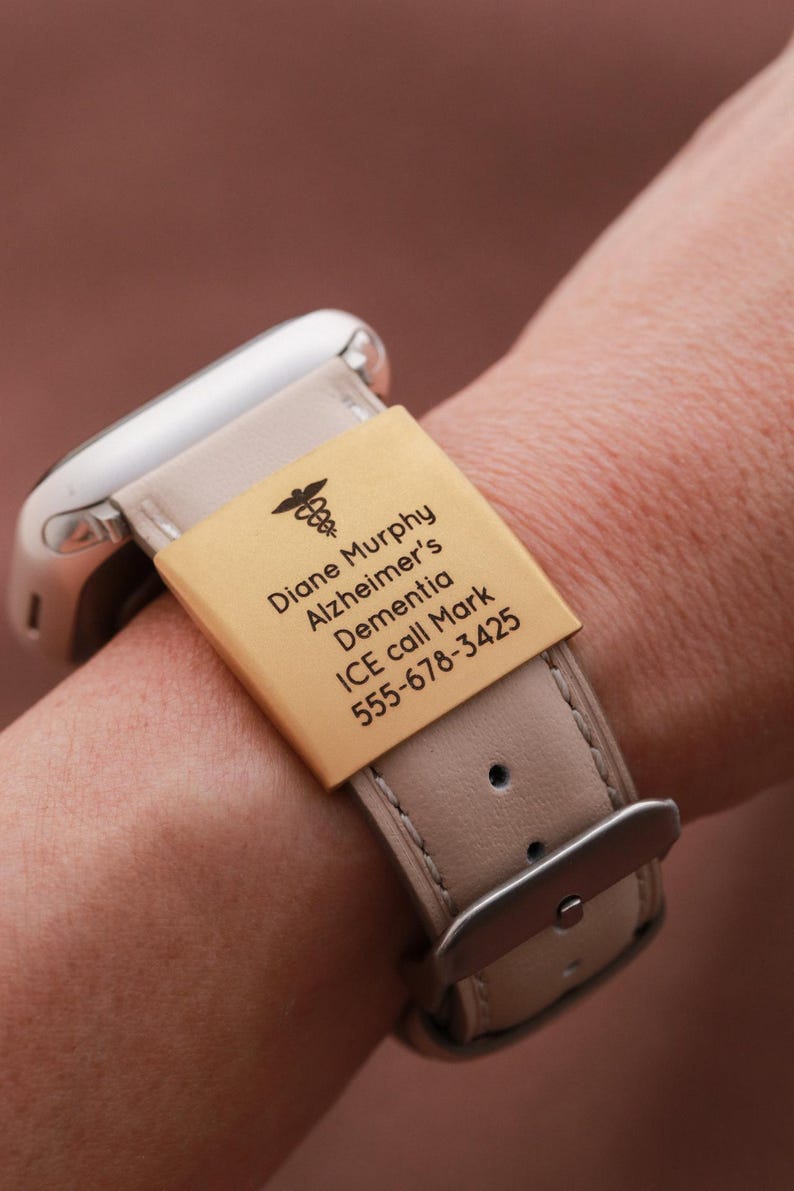 May include: A light beige watch band featuring a rectangular gold-toned metal medical alert tag engraved with a caduceus symbol and the text: Diane Murphy, Alzheimer's, Dementia, ICE call Mark, 555-678-3425. The band has a silver-toned buckle.