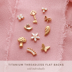 May include: A collection of gold-toned titanium threadless flat back studs featuring various shapes including a flower, key, bow, swan, wing, mushroom, butterfly, and a curved bar, all accented with clear crystals or smooth polished metal finishes.