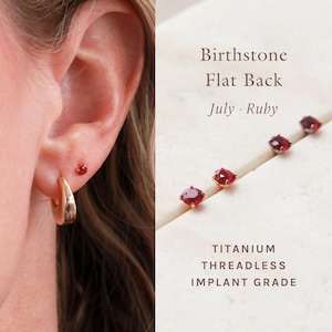 May include: Close-up of an ear with a gold hoop earring and a small red stud earring. The image also shows several small, red ruby birthstone earrings with gold settings. Text on the right reads "Birthstone Flat Back July - Ruby".