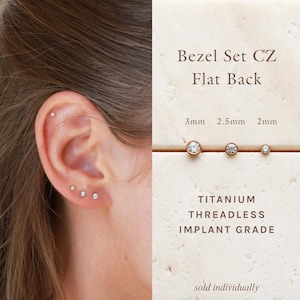 May include: Bezel set cubic zirconia flat back stud earrings in titanium. The image displays three sizes: 3mm, 2.5mm, and 2mm. The text reads Bezel Set CZ Flat Back, Titanium Threadless Implant Grade, and sold individually. The studs are shown in an ear.