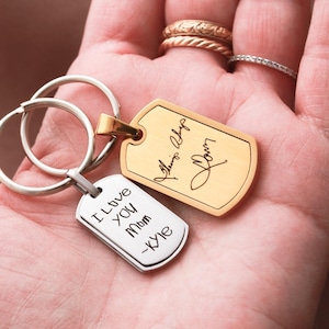May include: Two metal dog tag keychains with split rings. One tag is gold-toned with a cursive signature engraving. The other tag is silver-toned with the engraved text "I Love You Mom -Kyle" in a simple, handwritten-style font.