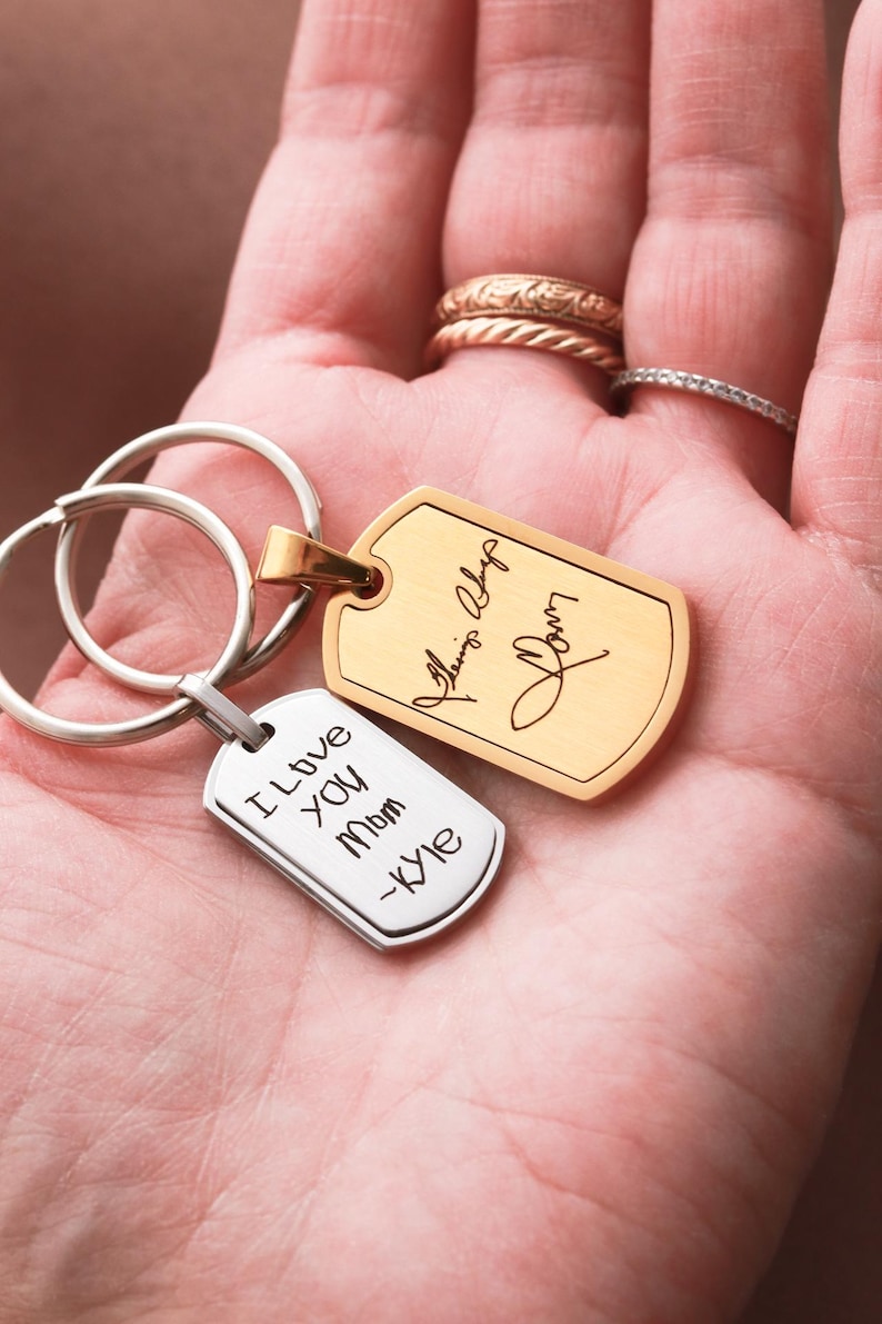 May include: Two metal dog tag keychains with split rings. One tag is gold-toned with a cursive signature engraving. The other tag is silver-toned with the engraved text "I Love You Mom -Kyle" in a simple, handwritten-style font.