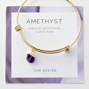 May include: Gold bangle bracelet with a small amethyst gemstone charm. The bracelet has a knotted closure and the charm is attached to the bracelet with a gold chain. The bracelet is displayed on a white card with the text "AMETHYST natural gemstone collection TOM DESIGN SHOP".