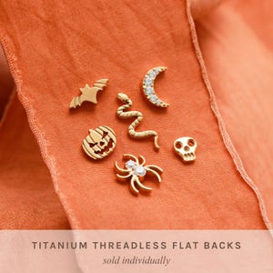May include: Gold-colored Halloween-themed titanium threadless flat back earrings. The collection includes a bat, snake, crescent moon, jack-o'-lantern, spider, and skull. The earrings are displayed on an orange fabric.