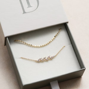 May include: Two gold necklaces in a white box. The top necklace is a simple chain with small round links. The bottom necklace has a delicate design of small leaves with clear stones.