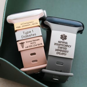 May include: Two silicone watch bands featuring medical alert tags. The pink band has gold, silver, and rose gold tags with text: allergies: egg, peanut, milk; Type 1 Diabetes; Emma Hansen, POTS, call 555-234-5678. The gray band has a silver tag with a medical symbol and text: ADRENAL INSUFFICIENCY, STEROID DEPENDANT, EPILEPSY.