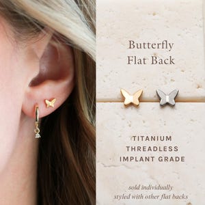 May include: A close-up of an ear with three earrings: a gold hoop with a dangling charm, and two butterfly-shaped studs. The text reads "Butterfly Flat Back" and "TITANIUM THREADLESS IMPLANT GRADE". One butterfly stud is gold, the other is silver.