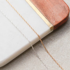 May include: Two delicate chains, one silver and one gold, lay side-by-side on a white marble surface. The chains are twisted and have a subtle shine.