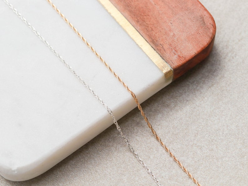 May include: Two delicate chains, one silver and one gold, lay side-by-side on a white marble surface. The chains are twisted and have a subtle shine.