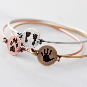 May include: Three thin metal bangles in silver, rose gold, and gold tones. Each bracelet features a small circular charm engraved with a dark image: one shows a paw print, one shows two baby footprints, and one shows a single small handprint.