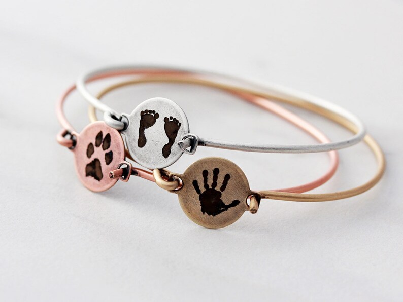 May include: Three thin metal bangles in silver, rose gold, and gold tones. Each bracelet features a small circular charm engraved with a dark image: one shows a paw print, one shows two baby footprints, and one shows a single small handprint.