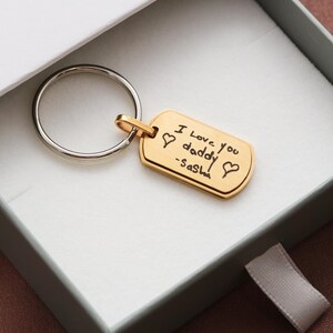 May include: A gold-toned rectangular metal keychain tag with rounded corners, attached to a silver-toned circular split ring. The tag features a black engraved inscription that reads I Love you daddy -Sasha, flanked by two small heart symbols.