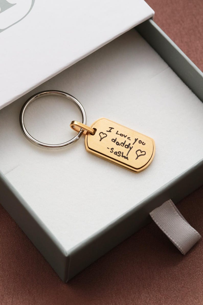 May include: A gold-toned rectangular metal keychain tag with rounded corners, attached to a silver-toned circular split ring. The tag features a black engraved inscription that reads I Love you daddy -Sasha, flanked by two small heart symbols.