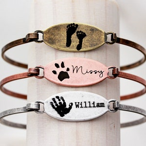 May include: Three oval-shaped metal bracelets in brass, copper, and silver tones. The top bracelet features two black footprints. The middle bracelet displays a black paw print and the name Missy. The bottom bracelet shows a black handprint and the name William.