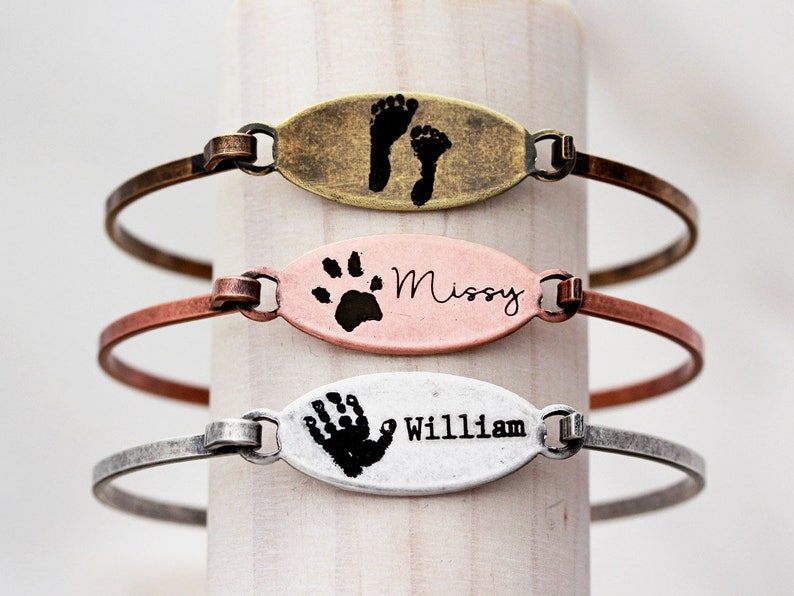May include: Three oval-shaped metal bracelets in brass, copper, and silver tones. The top bracelet features two black footprints. The middle bracelet displays a black paw print and the name Missy. The bottom bracelet shows a black handprint and the name William.