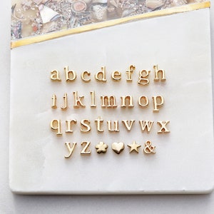 May include: Gold-toned alphabet letter charms and symbols, including a flower, heart, star, and ampersand. The charms are arranged in alphabetical order on a white surface.