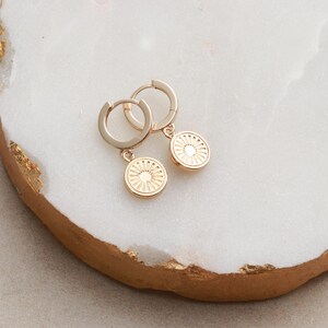 May include: A pair of gold hoop earrings with a small, circular pendant featuring a sunburst design.