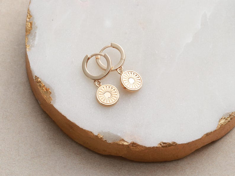 May include: A pair of gold hoop earrings with a small, circular pendant featuring a sunburst design.